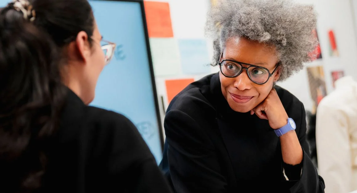 Staff member and student engaged in a conversation at The Manchester College, with classroom resources on the wall in the background.