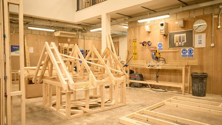 A carpentry workshop with timber roof frame models, workbenches, power tools, and measuring equipment used for construction training.