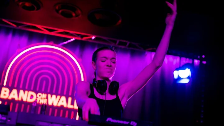 A DJ performing on stage at Band on the Wall, raising one arm while mixing music on the decks under vibrant purple lighting.