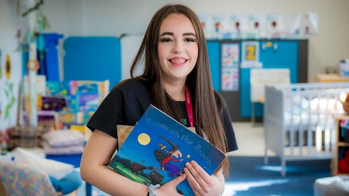 A learner in a childhood studies classroom holding children’s storybooks, standing in front of a colourful early years learning environment with books, toys, and activity stations.