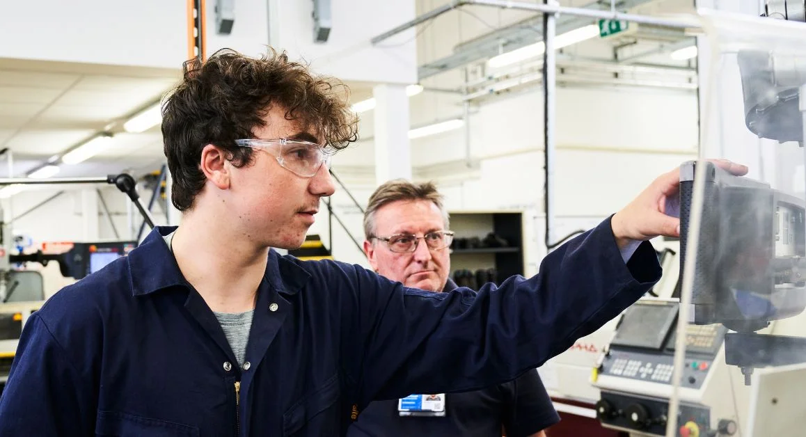 An apprentice operating machinery in an engineering workshop, with a Total People staff member observing.