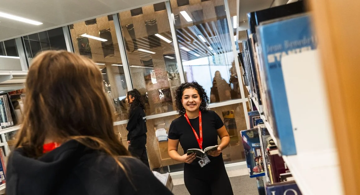 A student browsing books in The Manchester College library while holding an open book, with shelves and other students in the background.