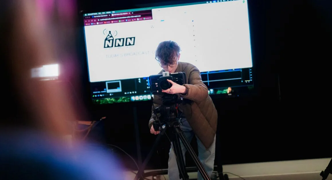 A media student from The Manchester College operating a professional video camera in a studio space, with a large screen displaying broadcast software behind.