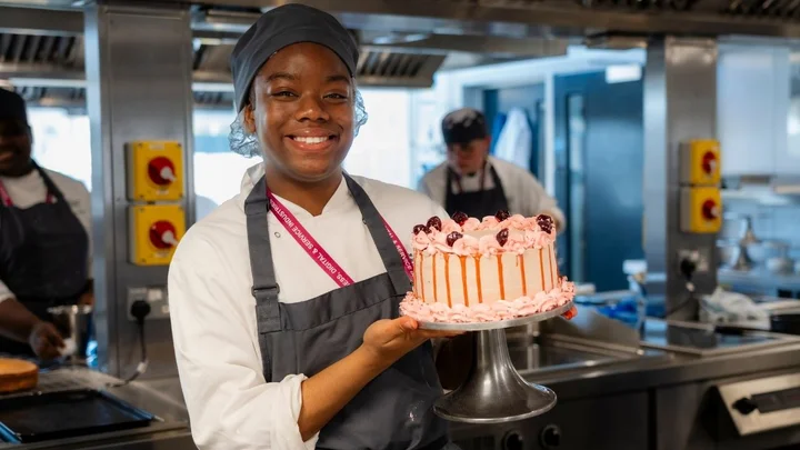 A catering learner holding a decorated pink cake in a professional training kitchen.