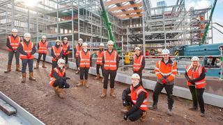 Construction students at the City Campus Manchester construction site, wearing high-visibility PPE and standing next to the steel structure of a new development.