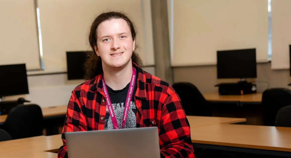 A student from The Manchester College wearing a lanyard sits in a computer room using a laptop, with several desks and computers visible behind them.