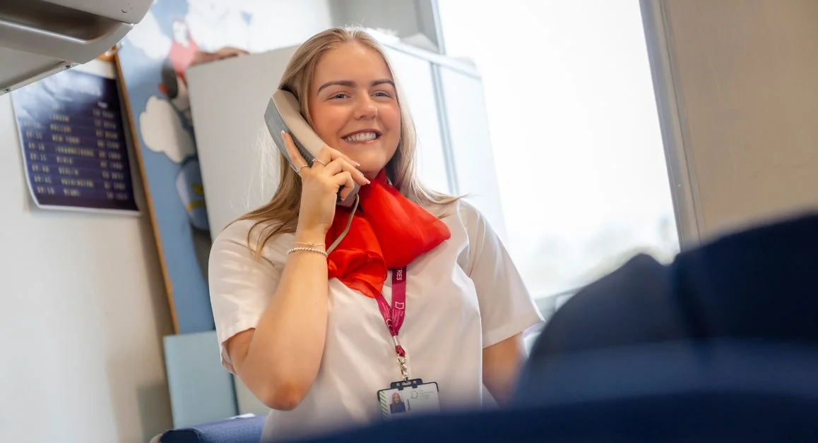 A learner from The Manchester College dressed in a uniform-style outfit speaking into a telephone handset during a practical training session, with classroom equipment visible behind.