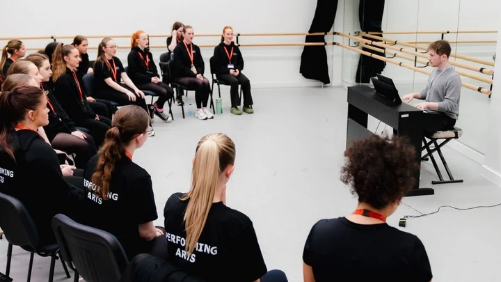 A performing arts class seated in a dance studio, taking part in a vocal warm‑up session led by an instructor at a keyboard.