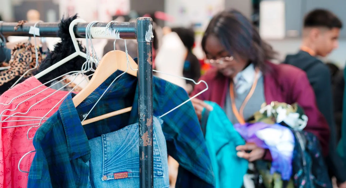 Clothing items hanging on a metal rail with people browsing garments in a busy indoor charity‑sale setting.