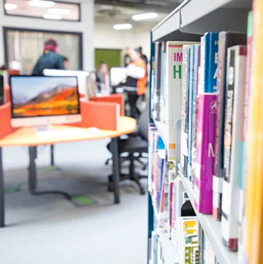 Library books on a shelf in the foreground, with a computer and study area blurred in the background.