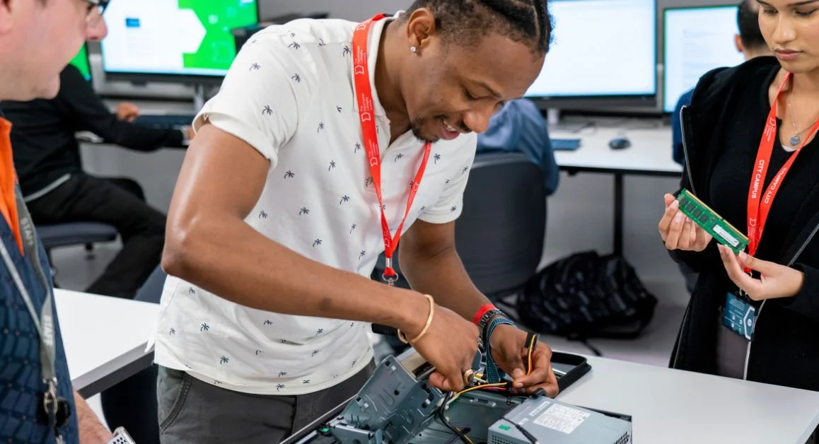 A computing learner working on the internal components of a desktop computer in a classroom at The Manchester College, with others observing and screens in the background.