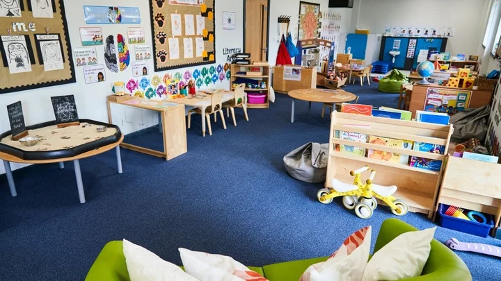A colourful early years classroom featuring activity stations, a reading corner, play tables, educational wall displays, and a wide range of learning resources designed for young children.