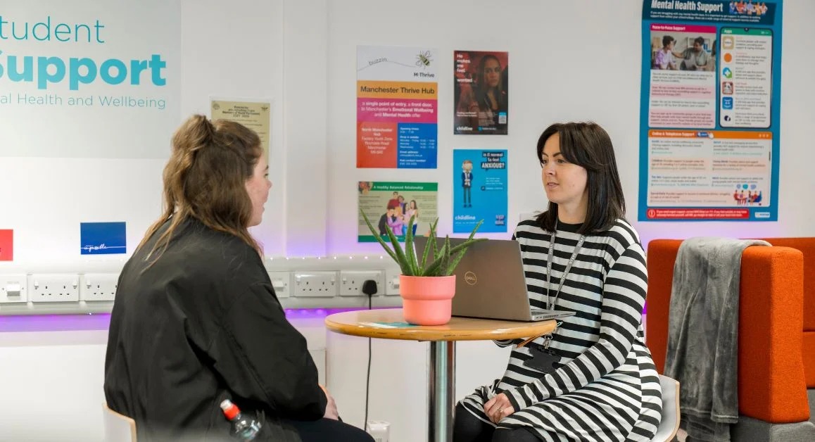 A pastoral support meeting taking place in a student support area, with two individuals seated at a small table beside wellbeing posters.