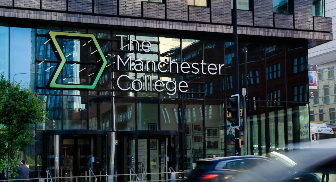 Exterior view of The Manchester College building, showing its glass entrance, signage, and surrounding city street with passing cars.