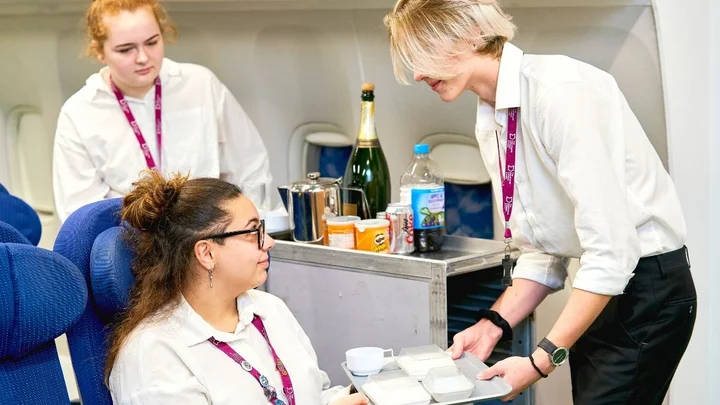 A travel and tourism learner serving an in‑flight meal to a seated passenger during a simulated cabin crew training session.
