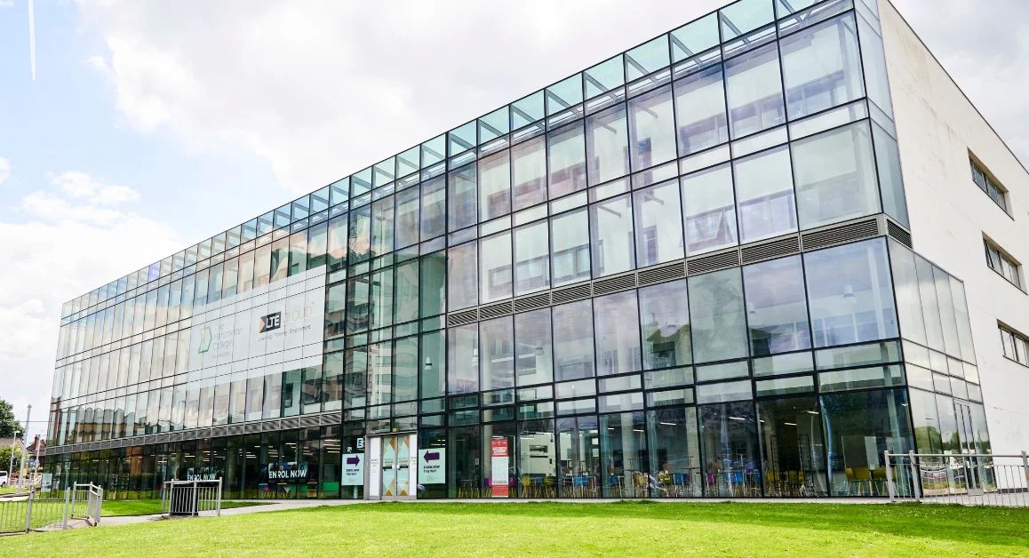Exterior of The Manchester College Wythenshawe Campus, a modern glass-fronted building with large windows and a green lawn in front.