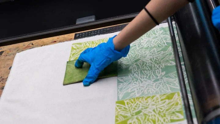 A learner printing a linocut design onto fabric using green ink and a hand roller during a printmaking session.