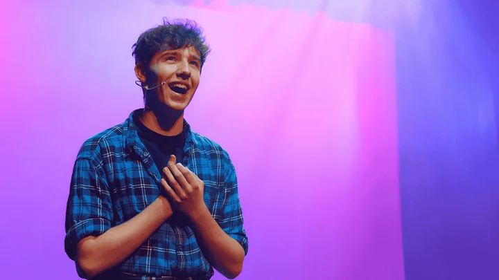 A performing arts learner standing on stage under colourful purple and blue lighting during a drama rehearsal.