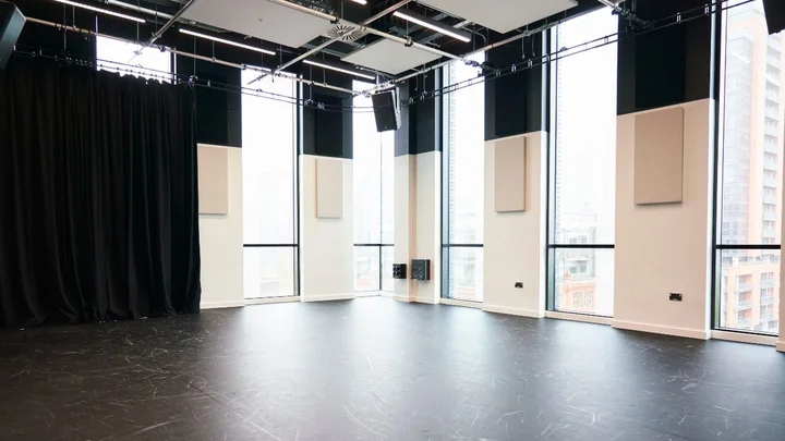 A large performing arts studio with full‑height windows, black flooring, wall‑mounted acoustic panels, and blackout curtains.