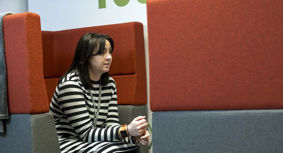 A person sitting in a high‑backed booth with red and grey padded panels inside a college student support hub talking to a student who is out of frame.