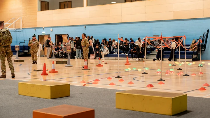 Public Services activity led by Army personnel inside a sports hall, featuring an obstacle course marked with cones and equipment while people observe from the seating area.