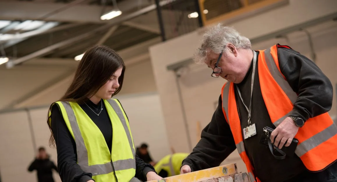 A learner from The Manchester College wearing a high‑visibility vest working with construction materials alongside a staff member in a workshop environment.