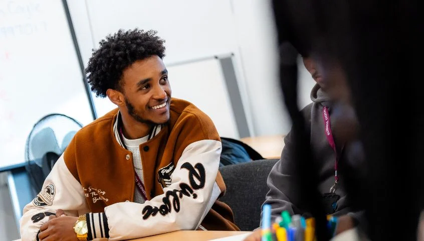 A learner sitting at a classroom table at The Manchester College, listening and smiling during a group discussion in a college setting.