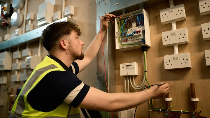 A learner in a workshop working on an electrical installation, testing wiring inside an open consumer unit while wearing a high‑visibility vest.