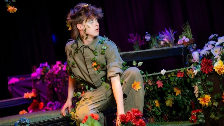 A performing arts learner on stage in a nature‑themed costume, surrounded by colourful flowers and foliage during a theatrical production.