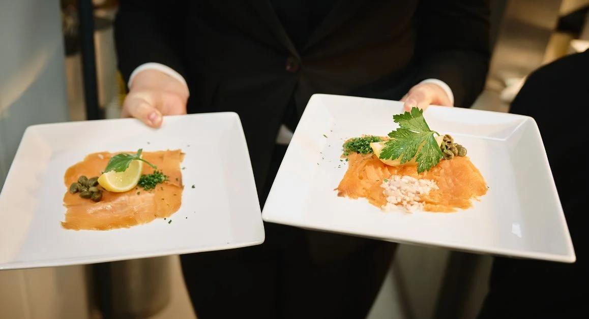 A server holding two square plates of smoked salmon in a professional training restaurant setting.