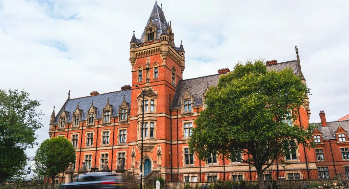 The Manchester College Nicholls Campus exterior view. Red-brick Victorian-style building and trees along the street.