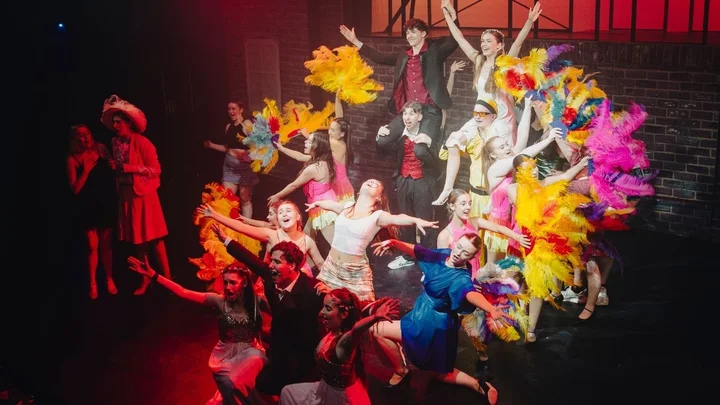 A performing arts ensemble on stage posing in a vibrant musical theatre finale, featuring colourful feather props and dramatic choreography under bright stage lighting.