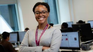Student wearing a lanyard in a modern computer lab, studying a T Level course at The Manchester College. Classmates working at computers in the background.