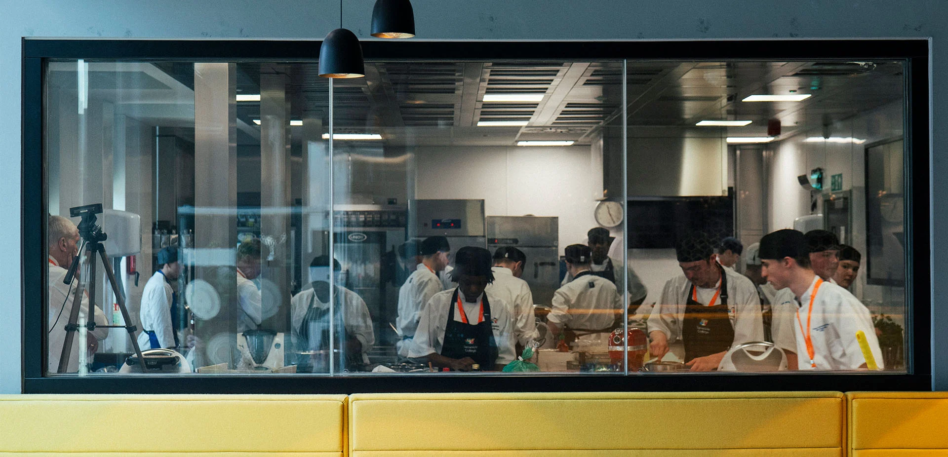 Learner kitchen at The Manchester College City Campus Manchester, showing people preparing food in a professional training kitchen viewed through a large observation window.”