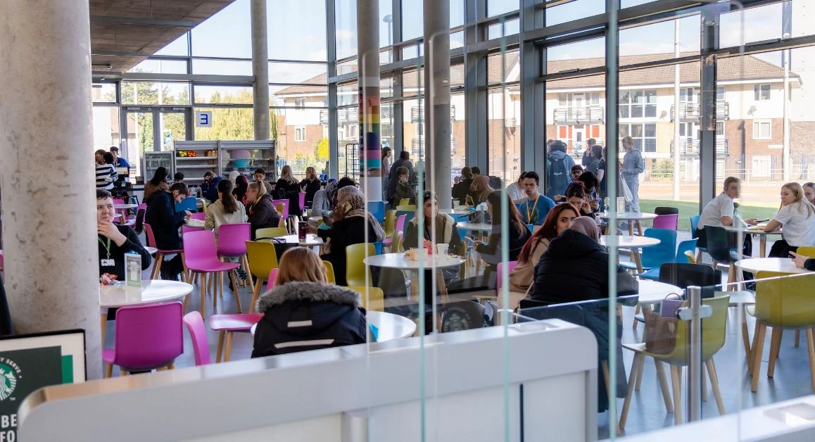 Students sitting at tables on colourful chairs in a bright, modern college café area, with large windows looking out onto buildings outside.