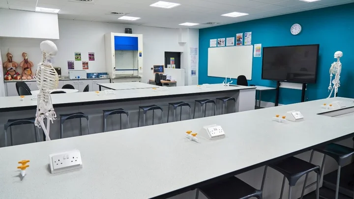 A modern science classroom with long laboratory benches, anatomical models, skeletons, a fume cupboard, and a large interactive screen.