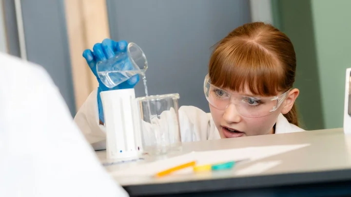 A science learner wearing safety goggles and gloves carefully pouring liquid from a beaker into a measuring cylinder during a laboratory experiment.