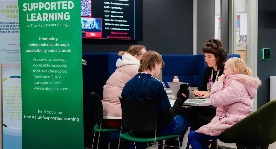 A Supported Learning information stand placed next to a group of people seated at a table for a meeting at The Manchester College.