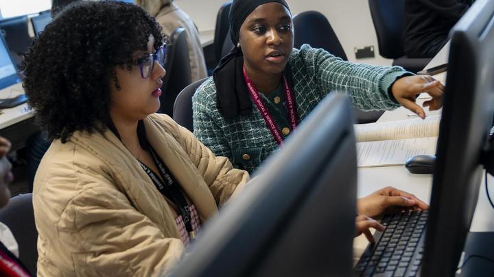 Two TMC students work together at a computer workstation in a classroom, with one student pointing at the screen while discussing work and the other using the keyboard.
