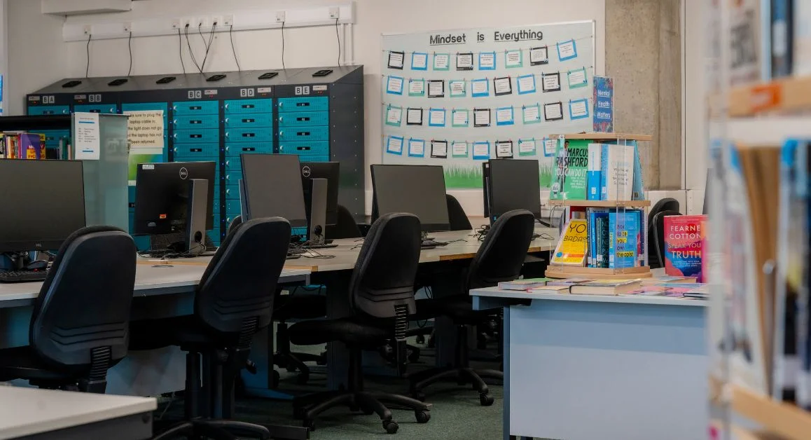 A computer suite with rows of desktop computers, chairs, bookshelves, and educational wall displays inside a study area at The Manchester College.