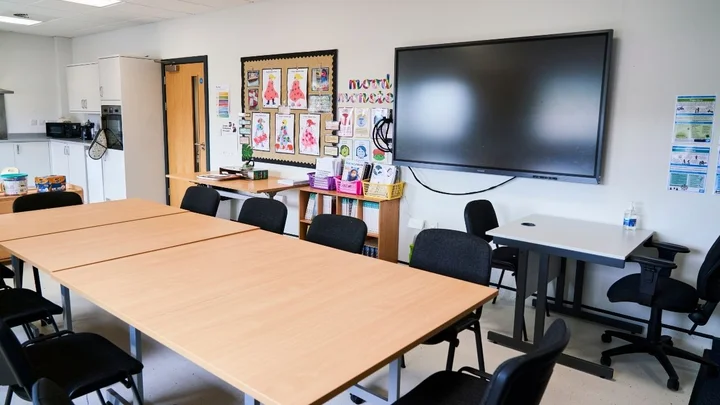 A classroom featuring collaborative tables, chairs, colourful wall displays, storage shelves with learning resources, and a large interactive screen.