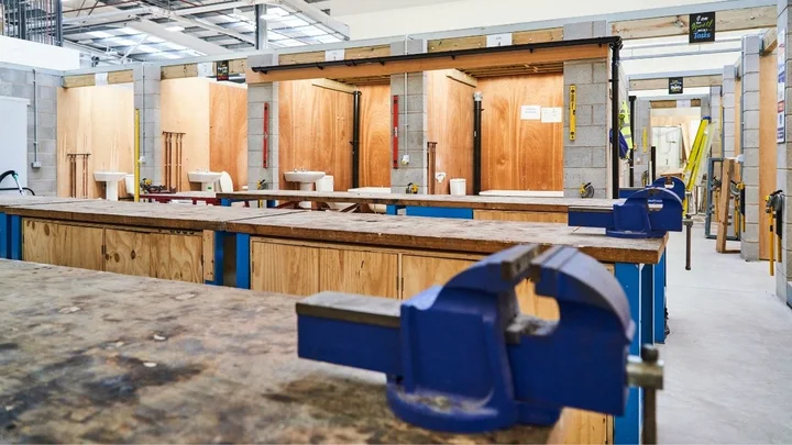 A plumbing and construction workshop featuring wooden practice bays, workbenches with mounted vices, and tools set up for hands‑on vocational training.