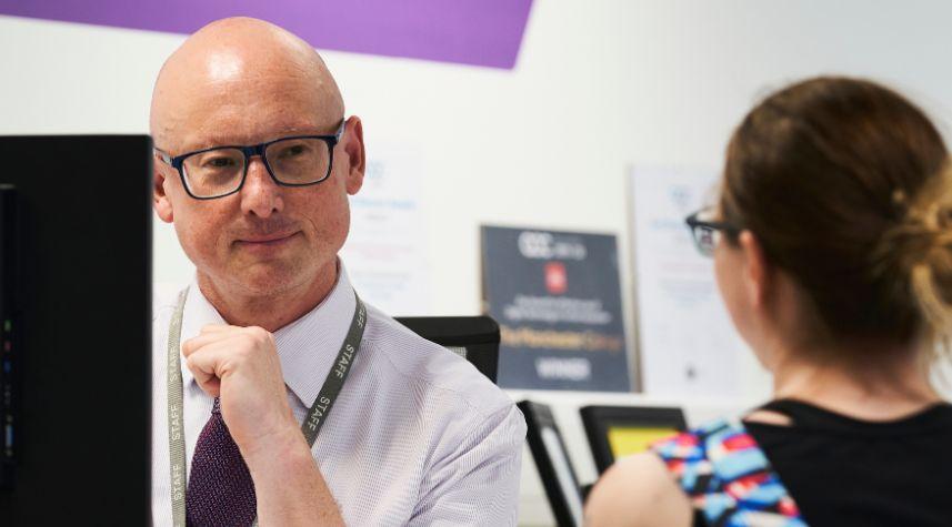 Staff member seated at a computer speaking with student who is partially out of frame.