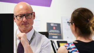 Staff member seated at a computer speaking with student who is partially out of frame.