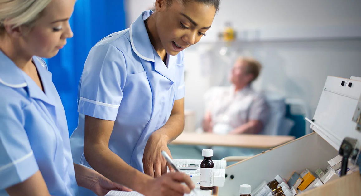Healthcare staff preparing medication from a medicine trolley, with a patient blurred in the background.