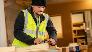 Person wearing a high-visibility vest uses a hand plane to smooth a piece of timber in a construction workshop, with woodworking equipment and materials visible in the background.