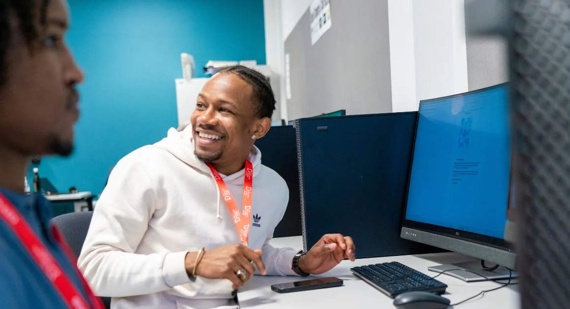 A learner from The Manchester College sitting at a computer desk in a digital skills classroom, smiling while speaking with another student.