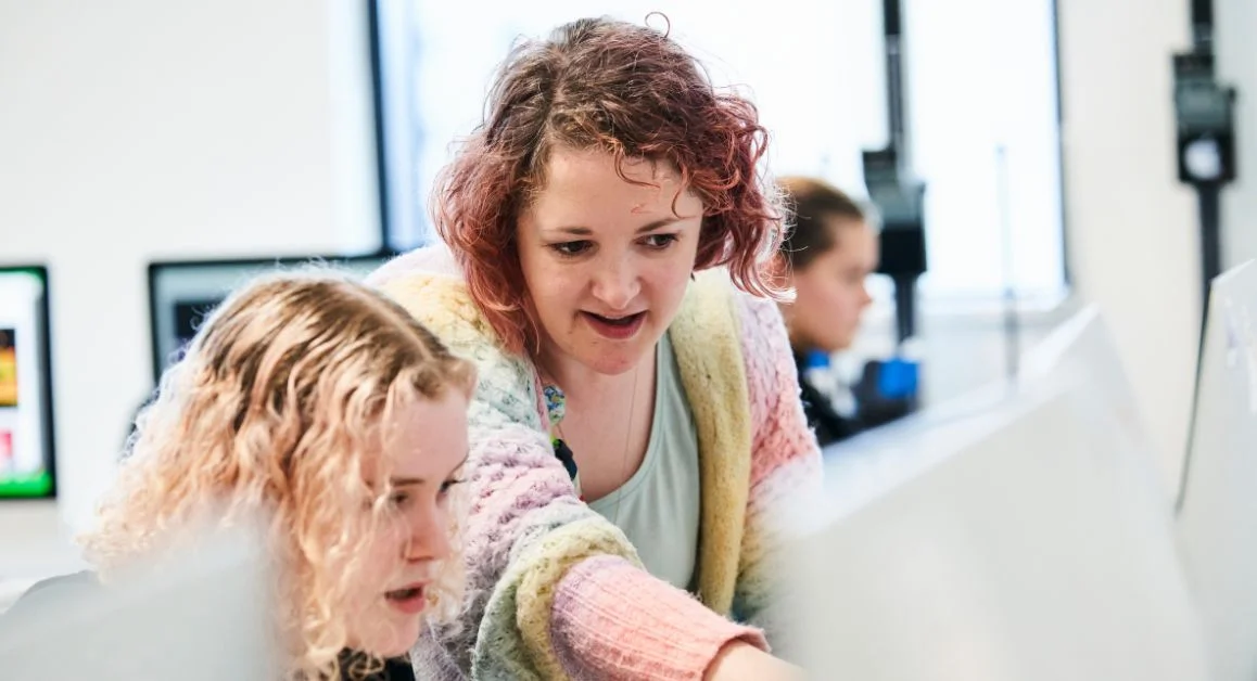 A tutor assisting a student at The Manchester College, pointing at a screen, with bright classroom lighting and multiple monitors visible.