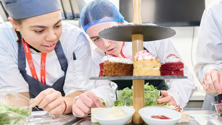 Catering learners preparing dishes with a tiered cake stand and food ingredients arranged on the workstation.