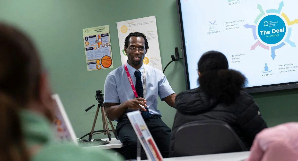 A speaker presents to a small group in a classroom, with a presentation screen visible in the background.